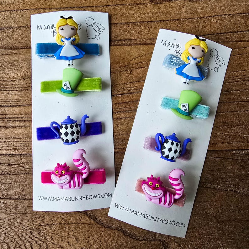 Op de afbeelding: Haarclips met Alice in Wonderland-ontwerpen. Elke clip heeft een ander ontwerp: Alice, de hoed van de Mad Hatter, een theepot en de Cheshire Cat. De clips zitten op witte kaarten met de website www.mamabunnybows.com.