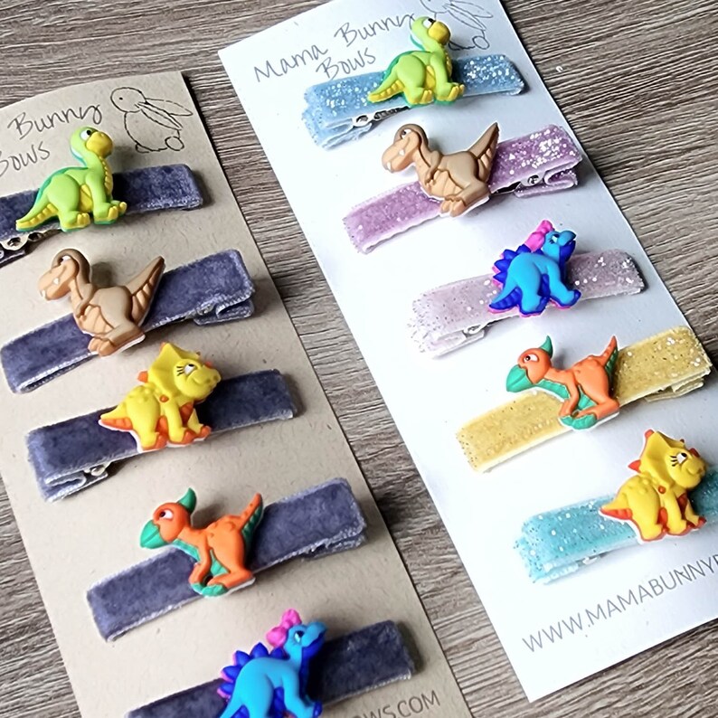 May include: A set of 10 dinosaur hair clips on velvet and glitter backing. The clips feature a variety of dinosaurs in different colors, including green, orange, blue, and brown. The text "Mama Bunny Bows" and "www.mamabunnybows.com" are visible on the backing.