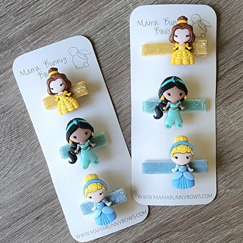 May include: Three sets of princess-themed hair clips. Each set includes a gold, blue, or green glitter barrette with a princess character on top. The princesses are Belle, Jasmine, and Cinderella. The packaging includes the text "Mama Bunny Bows" and a website address.