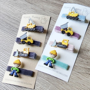 May include: A set of 7 construction vehicle hair clips on a card with the text "Mama Bunny Bows" and a website address. The clips feature a variety of construction vehicles, including a crane, dump truck, bulldozer, and a construction worker.