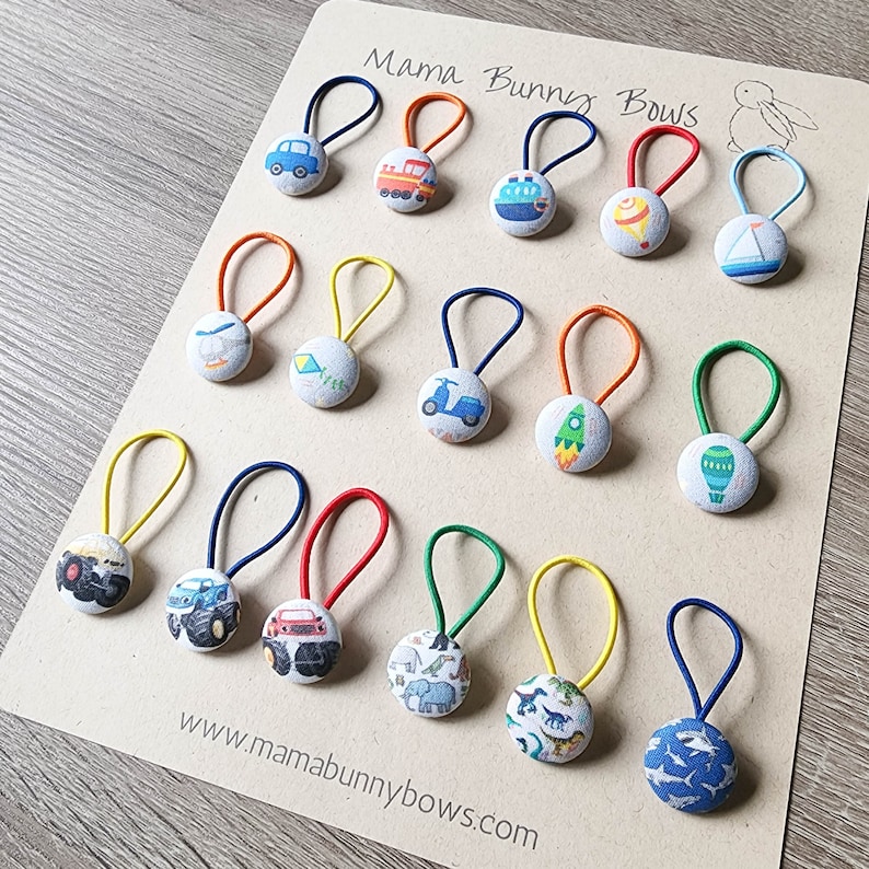 May include: A set of 18 colorful hair ties with various fabric buttons featuring different designs, including cars, airplanes, rockets, and dinosaurs. The hair ties are displayed on a white card with the text "Mama Bunny Bows" and a website address.