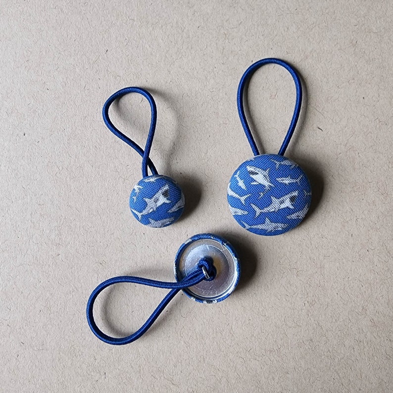 May include: Two blue hair ties with white shark print fabric. One hair tie is larger than the other. The larger hair tie has a button-like closure. The smaller hair tie has a simple loop closure.