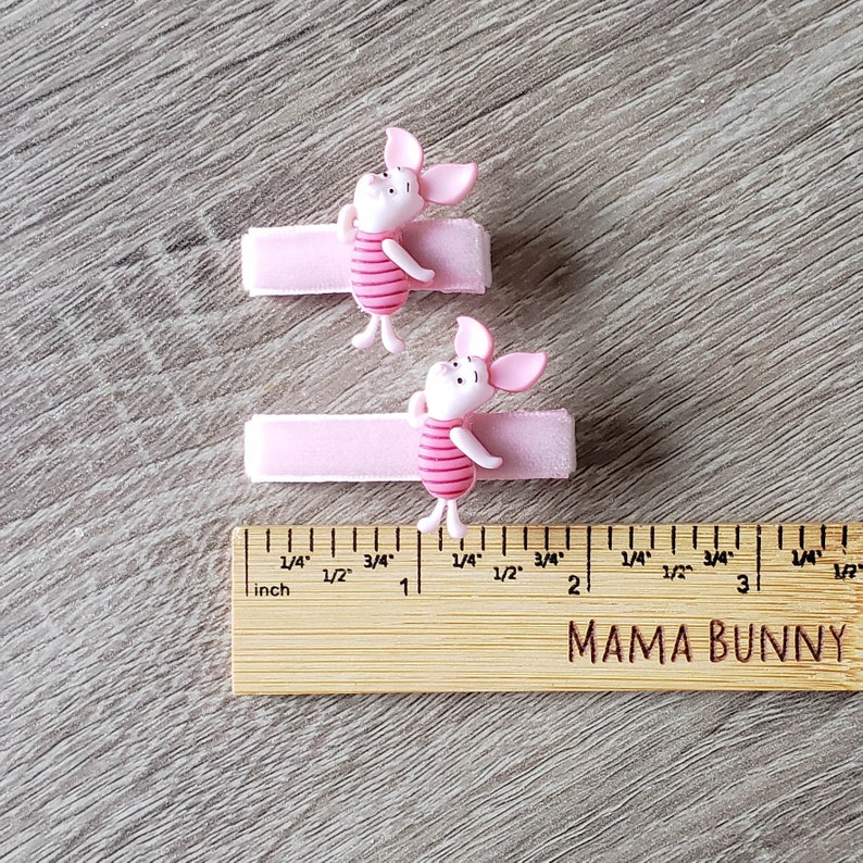 Puede incluir: Dos pinzas para el pelo de cerdo rosa con un dise&ntilde;o de cerdo de dibujos animados. El cerdo lleva una camisa a rayas rosa y blanca. Las pinzas miden 2,5 cm de largo y se muestran junto a una regla.