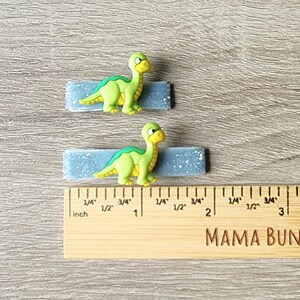May include: Two blue glitter hair clips with green dinosaur figures attached. The clips are approximately 1 inch long. The ruler in the image is marked in inches.