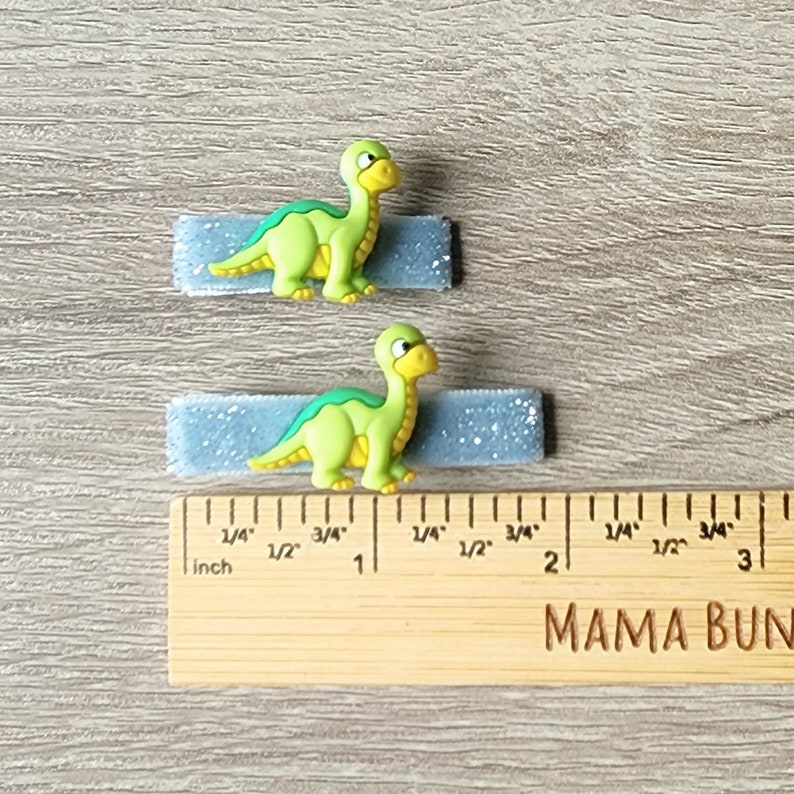 May include: Two blue glitter hair clips with green dinosaur figures attached. The clips are approximately 1 inch long. The ruler in the image is marked in inches.
