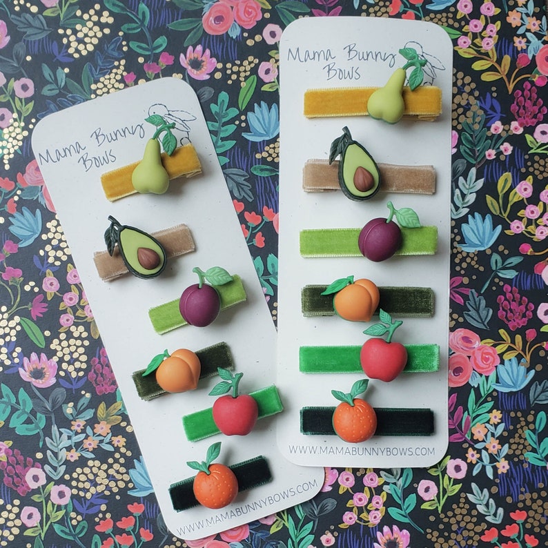 K&ouml;nnte beinhalten: Zwei Karten mit dem Text "Mama Bunny Bows" und einer Website-Adresse. Jede Karte enth&auml;lt eine Reihe von Haarklammern mit verschiedenen Fruchtmotiven, darunter eine Birne, eine Avocado, eine Pflaume, ein Pfirsich, ein Apfel und eine Orange.