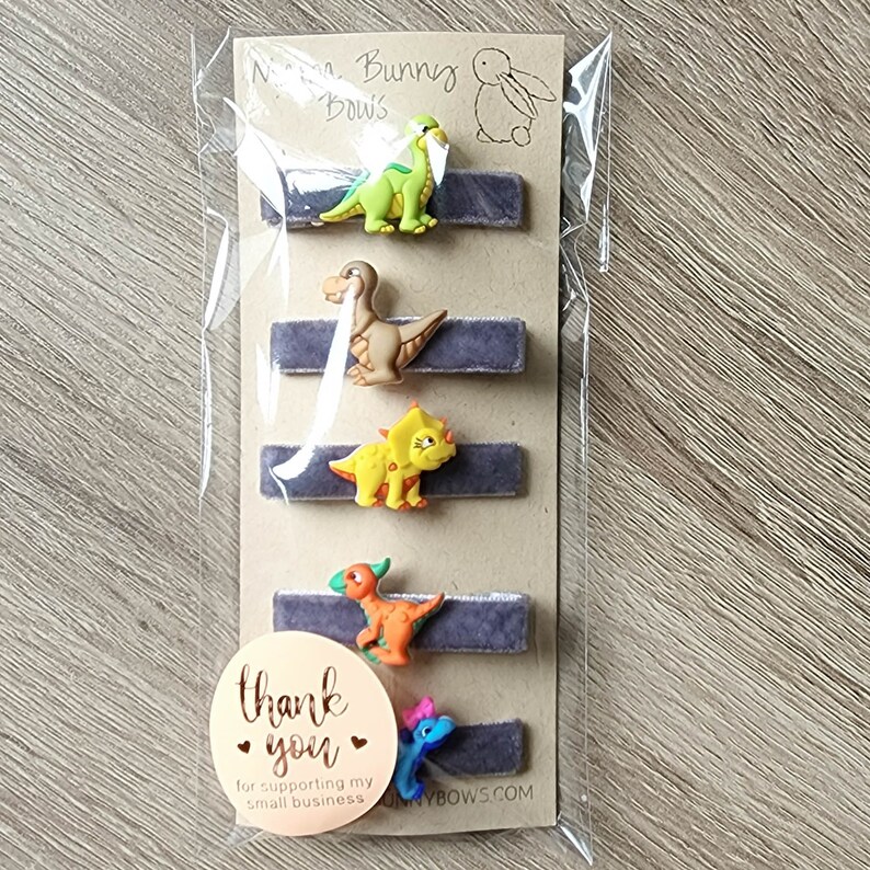 May include: Set of five grey velvet hair clips with dinosaur charms. The dinosaurs are green, brown, yellow, orange, and blue. The packaging includes a thank you sticker that says "Thank you for supporting my small business".