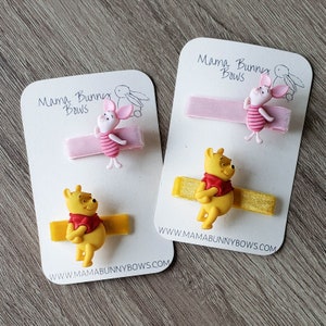 Puede incluir: Dos juegos de pinzas para el pelo con los personajes de Winnie de Pooh y Piglet. Cada juego incluye una cinta amarilla y rosa con un amuleto de personaje a juego. Las tarjetas tienen el texto "Mama Bunny Bows" y una direcci&oacute;n de sitio web.