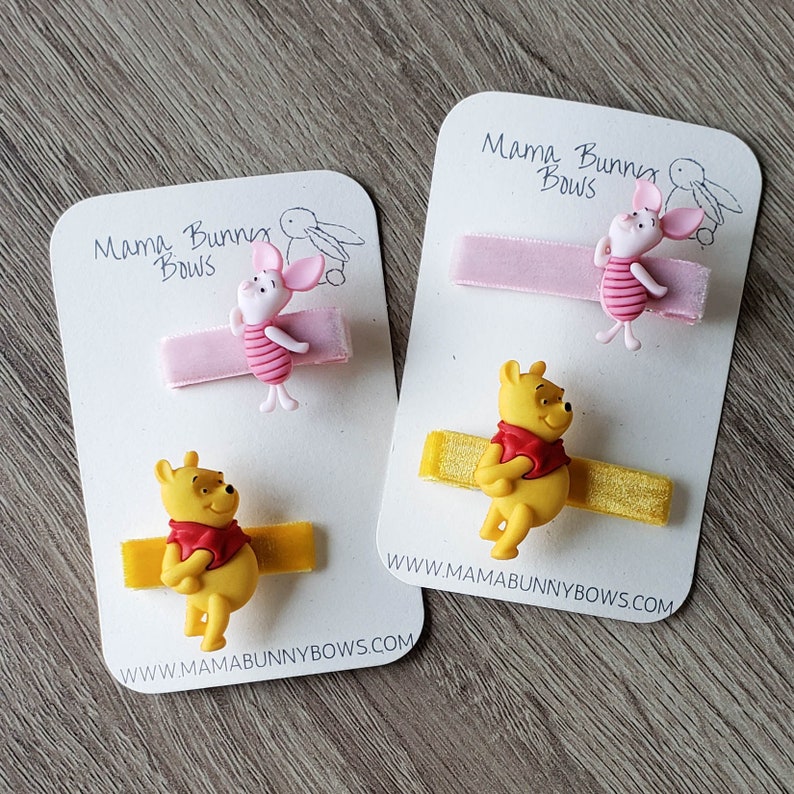 Puede incluir: Dos juegos de pinzas para el pelo con los personajes de Winnie de Pooh y Piglet. Cada juego incluye una cinta amarilla y rosa con un amuleto de personaje a juego. Las tarjetas tienen el texto "Mama Bunny Bows" y una direcci&oacute;n de sitio web.