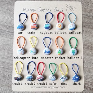 May include: A set of 18 colorful hair ties with fabric covered buttons featuring various transportation and animal themes. The buttons include a car, train, tugboat, balloon, sailboat, helicopter, kite, scooter, rocket, balloon, truck, monster truck, safari animals, dinosaur, and shark. The hair ties are displayed on a wooden background with the text "Mama Bunny Bows" and "www.mamabunnybows.com".