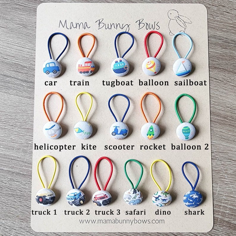 May include: A set of 18 colorful hair ties with fabric covered buttons featuring various transportation and animal themes. The buttons include a car, train, tugboat, balloon, sailboat, helicopter, kite, scooter, rocket, balloon, truck, monster truck, safari animals, dinosaur, and shark. The hair ties are displayed on a wooden background with the text "Mama Bunny Bows" and "www.mamabunnybows.com".