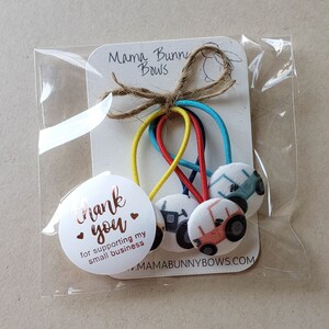 May include: Set of five colorful hair ties with tractor fabric buttons. The hair ties are packaged in a clear plastic bag with a white card that says "Mama Bunny Bows" and "Thank you for supporting my small business" with a heart. The card also has the website address www.mamabunnybows.com.