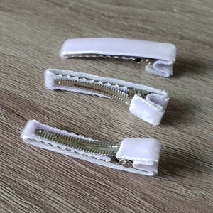 May include: Three white velvet hair clips with silver metal clasps. The clips are arranged in a row, with the top clip being the largest and the bottom clip being the smallest.