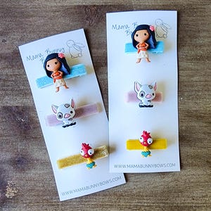 May include: Three sets of hair clips featuring characters from the animated movie Moana. The first set includes a blue clip with Moana, a pink clip with Pua the pig, and a gold clip with Hei Hei the rooster. The second set includes a blue clip with Moana, a pink clip with Pua the pig, and a yellow clip with Hei Hei the rooster. The cards are white with the text "Mama Bunny Bows" and the website address "www.mamabunnybows.com".
