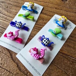 Op de afbeelding: Haarclips met Alice in Wonderland-personages. Elke clip heeft een ander ontwerp, waaronder Alice, een hoed van de Mad Hatter, een theepot en de Cheshire Cat. De clips zijn bevestigd aan witte kaarten met de tekst "MAMABUNNYBOWS.COM".
