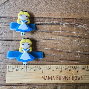 Op de afbeelding: Twee haarspeldjes met Alice in Wonderland-karakter. Elk speldje heeft een blondharige Alice-figuur in een blauwe jurk en een zwarte strik. De speldjes zijn bevestigd aan een blauw lint. Een houten liniaal met de tekst "MAMA BUNNY BOWS" staat in de afbeelding.