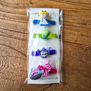 Op de afbeelding: Vier haarclips met Alice in Wonderland-thema. De clips tonen Alice, de hoed van de Mad Hatter, een theepot en de Cheshire Cat. Elk personage is bevestigd aan een gekleurd fluwelen lint. Het pakket bevat een bedanksticker.