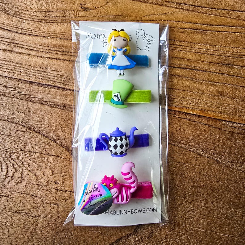 Op de afbeelding: Vier haarclips met Alice in Wonderland-thema. De clips tonen Alice, de hoed van de Mad Hatter, een theepot en de Cheshire Cat. Elk personage is bevestigd aan een gekleurd fluwelen lint. Het pakket bevat een bedanksticker.