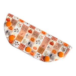 May include: A semi-circular fabric mask extender featuring a patchwork pattern of pumpkins, autumn leaves, and brown plaid squares. The accessory includes five bright orange plastic snap buttons arranged along the curved edge for adjustment.