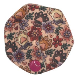 May include: A pink, floral-patterned pet accessory with paw prints and the text "THE BEANS" on banners. The accessory has a pink button closure. The design includes flowers in shades of red, purple, and pink.
