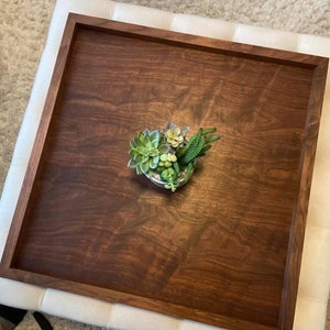 May include: A square, dark brown wooden tray with a raised border. The tray holds a small arrangement of succulents in a decorative container. The tray is placed on a white upholstered ottoman.