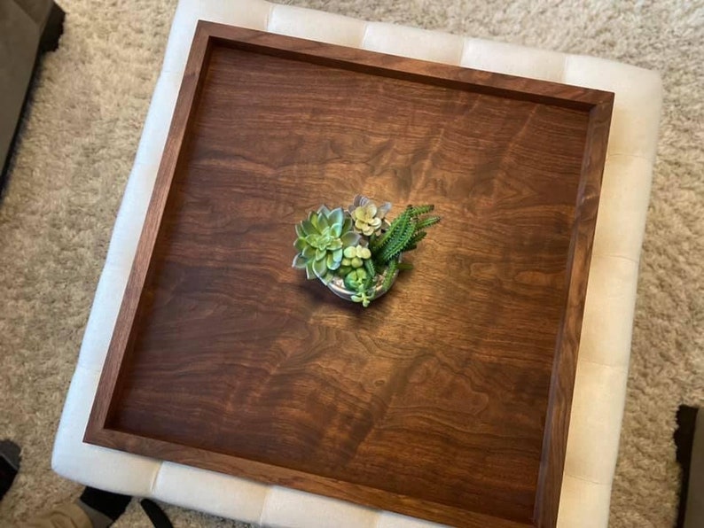 May include: A square, dark brown wooden tray with a raised border. The tray holds a small arrangement of succulents in a decorative container. The tray is placed on a white upholstered ottoman.