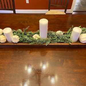May include: A wooden rectangular tray with a rustic brown finish, filled with green eucalyptus sprigs, white pumpkins, and three white pillar candles. The tray is sitting on a dark wood table.