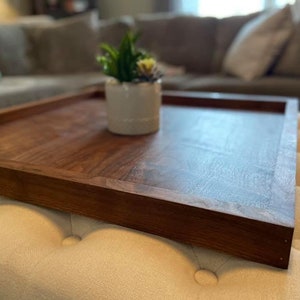 May include: A rectangular wooden serving tray with a rich, dark brown finish. The tray has raised edges and is placed on a cream-colored ottoman. A small white ceramic pot with a succulent plant sits on the tray.