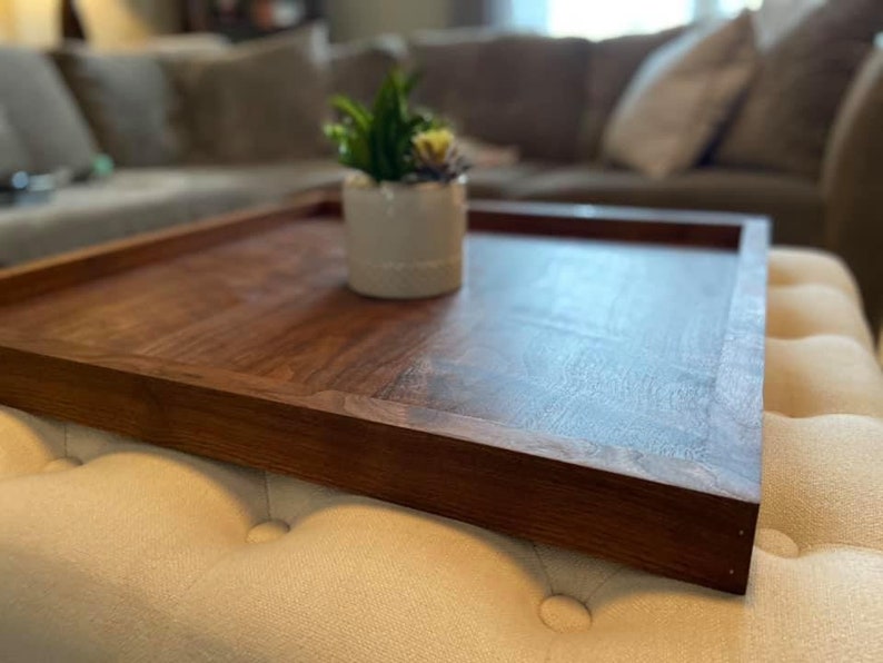 May include: A rectangular wooden serving tray with a rich, dark brown finish. The tray has raised edges and is placed on a cream-colored ottoman. A small white ceramic pot with a succulent plant sits on the tray.