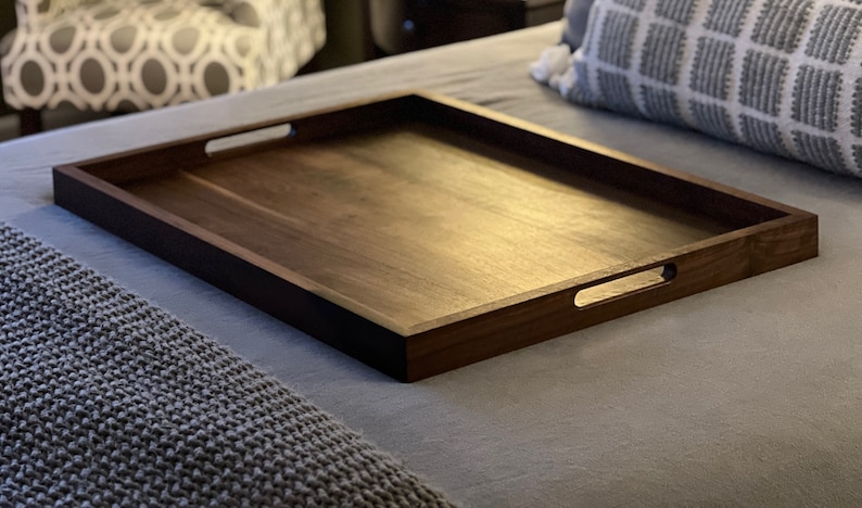 May include: A rectangular wooden serving tray with a dark brown finish. The tray has cut-out handles on each side. The tray is placed on a bed with a gray blanket and pillow.