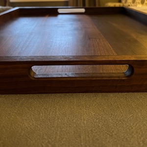 May include: A rectangular wooden serving tray with a rich, dark brown finish. The tray features integrated handles on each side, designed for easy carrying. The wood grain is visible, adding to the tray's natural aesthetic. This tray is suitable for serving food and drinks.
