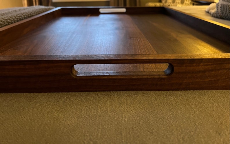 May include: A rectangular wooden serving tray with a rich, dark brown finish. The tray features integrated handles on each side, designed for easy carrying. The wood grain is visible, adding to the tray's natural aesthetic. This tray is suitable for serving food and drinks.