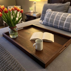 May include: A rectangular wooden serving tray with a dark brown finish, featuring built-in handles. A white mug and an open book rest on the tray. A glass jar with a leather handle holds a bouquet of red and yellow tulips. The tray is on a bed.