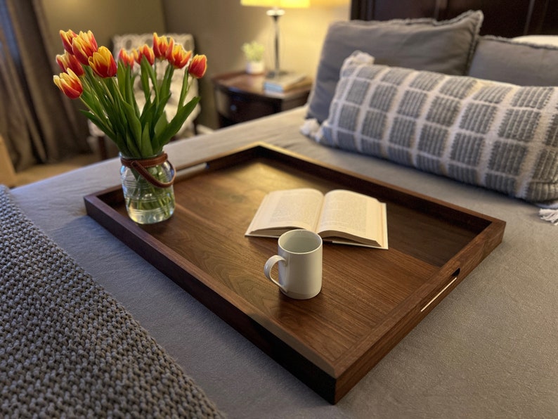 May include: A rectangular wooden serving tray with a dark brown finish, featuring built-in handles. A white mug and an open book rest on the tray. A glass jar with a leather handle holds a bouquet of red and yellow tulips. The tray is on a bed.