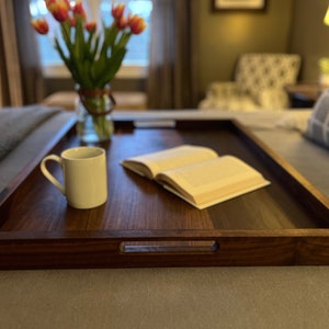 May include: A rectangular, dark wood serving tray with integrated handles. A white mug and an open book rest on the tray. A vase of tulips sits in the background, suggesting a cozy, inviting setting. The tray is on a bed.