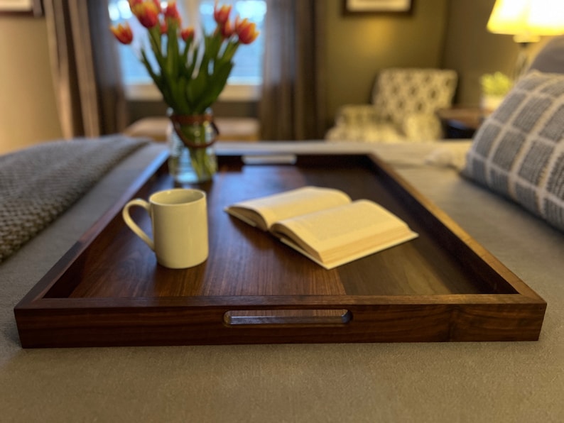 May include: A rectangular, dark wood serving tray with integrated handles. A white mug and an open book rest on the tray. A vase of tulips sits in the background, suggesting a cozy, inviting setting. The tray is on a bed.