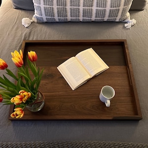May include: A wooden serving tray with a dark brown finish holds a vase of yellow and red tulips, an open book, and a white mug. The tray is on a gray bed with a decorative pillow.
