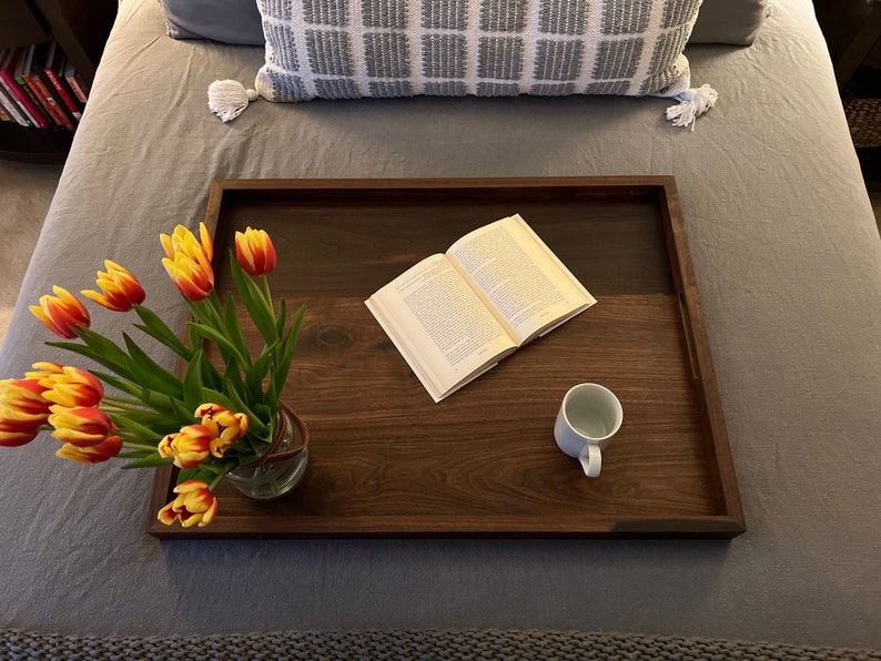 May include: A wooden serving tray with a dark brown finish holds a vase of yellow and red tulips, an open book, and a white mug. The tray is on a gray bed with a decorative pillow.