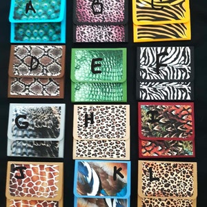 May include: A collection of small, rectangular wallets with various animal print designs. The wallets feature patterns such as peacock feathers, leopard spots, zebra stripes, and snake scales. Each wallet has a contrasting colored border and a flap closure.