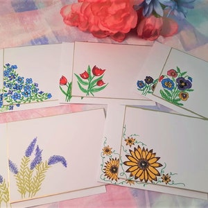 May include: A collection of white stationery cards with hand-painted floral designs. Each card features a different flower arrangement, including blue forget-me-nots, red tulips, colorful pansies, lavender, and a yellow sunflower. The cards have gold borders.