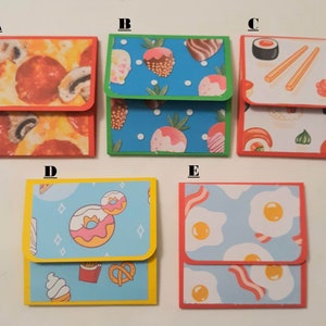 May include: A collection of five small, square paper wallets with colorful food-themed designs. The designs include pizza, sushi, donuts, fruit, and eggs with bacon. Each wallet has a contrasting border color.