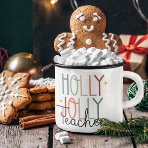 May include: White enamel mug with the text "Holly Jolly Teacher" in colorful letters. The mug is filled with marshmallows and topped with a gingerbread man cookie.