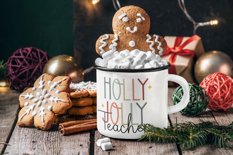 May include: White enamel mug with the text "Holly Jolly Teacher" in colorful letters. The mug is filled with marshmallows and topped with a gingerbread man cookie.