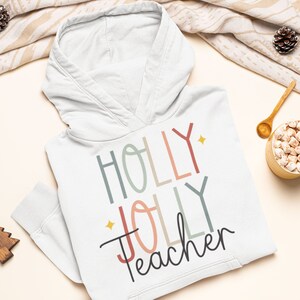 May include: White hooded sweatshirt with the text "HOLLY JOLLY Teacher" in colorful, stylized lettering. The text is in a rainbow of colors, including red, pink, blue, green, and yellow. The sweatshirt is laid flat on a white surface with a cup of hot chocolate and other holiday decorations.