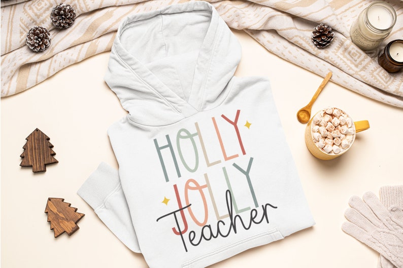 May include: White hooded sweatshirt with the text "HOLLY JOLLY Teacher" in colorful, stylized lettering. The text is in a rainbow of colors, including red, pink, blue, green, and yellow. The sweatshirt is laid flat on a white surface with a cup of hot chocolate and other holiday decorations.