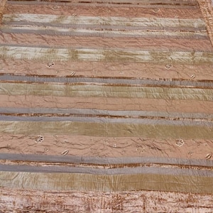 May include: A large, rectangular quilt with horizontal stripes in shades of gold, brown, and gray. The quilt has a textured appearance and is embellished with small, embroidered floral designs. The edges are finished with a ruffled border.