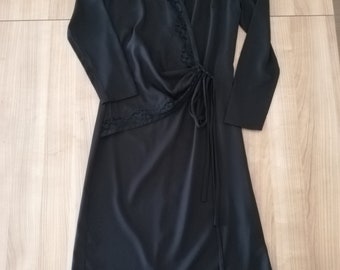 May include: A black wrap dress with long sleeves and a V-neckline. The dress features a tie closure at the waist and delicate lace trim along the neckline and sleeve cuffs. The dress is made of a smooth, flowing fabric.