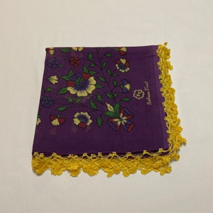 May include: A purple handkerchief with a floral pattern in red, green, blue, and yellow. The handkerchief has a yellow crocheted lace border and the text "Selma Firat".
