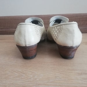May include: Pair of off-white leather shoes with woven detailing on the sides. The shoes have a low, stacked heel with a dark brown finish. The shoes are set against a light brown wooden surface.