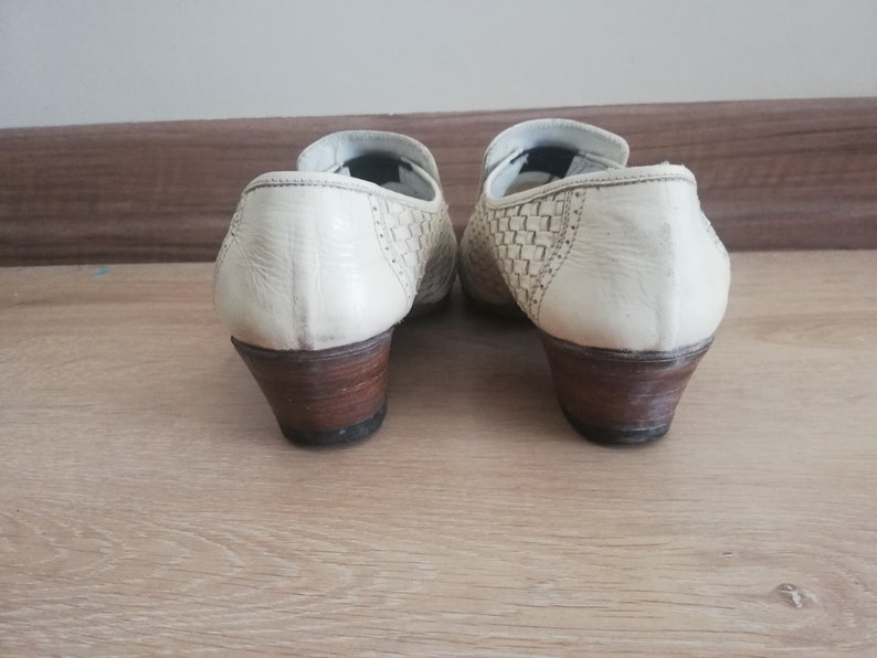 May include: Pair of off-white leather shoes with woven detailing on the sides. The shoes have a low, stacked heel with a dark brown finish. The shoes are set against a light brown wooden surface.